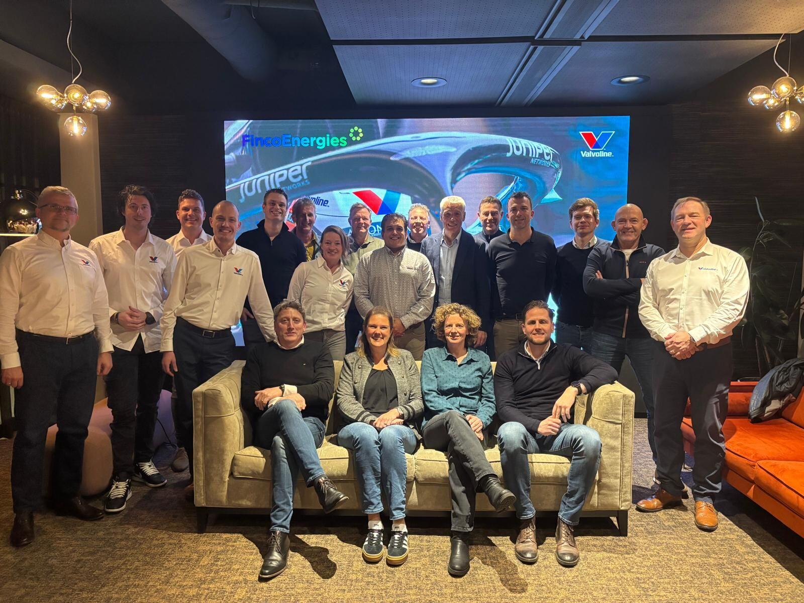 Group photo of a team of employees from FincoEnergies and Valvoline, posing in an informal setting with a large screen in the background displaying the logos of both companies.